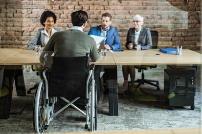 Rückansicht eines männlichen Bewerbers im Rollstuhl, der dem Personalteam seine Bewerbungsunterlagen für das Vorstellungsgespräch überreicht.