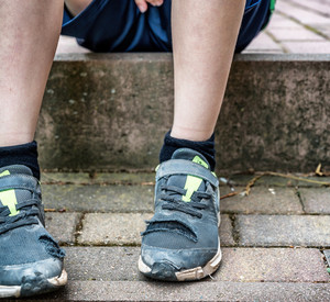Unkenntliches Kind sitzt mit kaputten Schuhen auf Gehweg