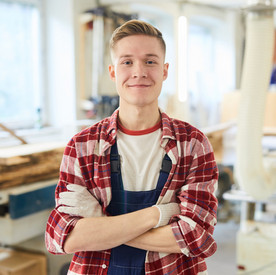 Ein Zimmerei-Student steht mit Arbeitshandschuhen in einer Werkstatt und schaut frontal in die Kamera. 